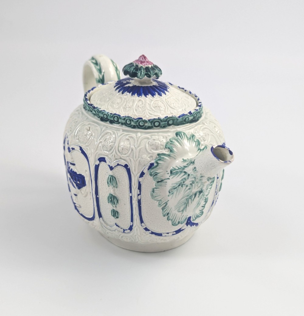 An 18th-century lead-glazed ceramic teapot featuring relief-moulded arches, blue and green painted accents, and a moulded lid with a pink flower finial. View V
