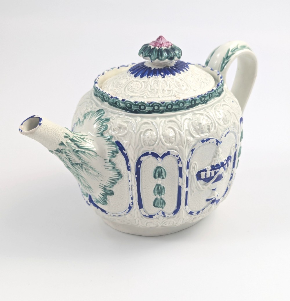 An 18th-century lead-glazed ceramic teapot featuring relief-moulded arches, blue and green painted accents, and a moulded lid with a pink flower finial. View VI