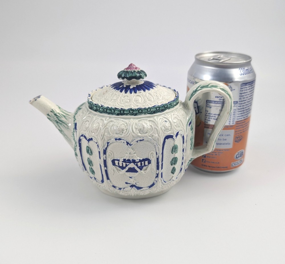 An 18th-century lead-glazed ceramic teapot featuring relief-moulded arches, blue and green painted accents, and a moulded lid with a pink flower finial. View X