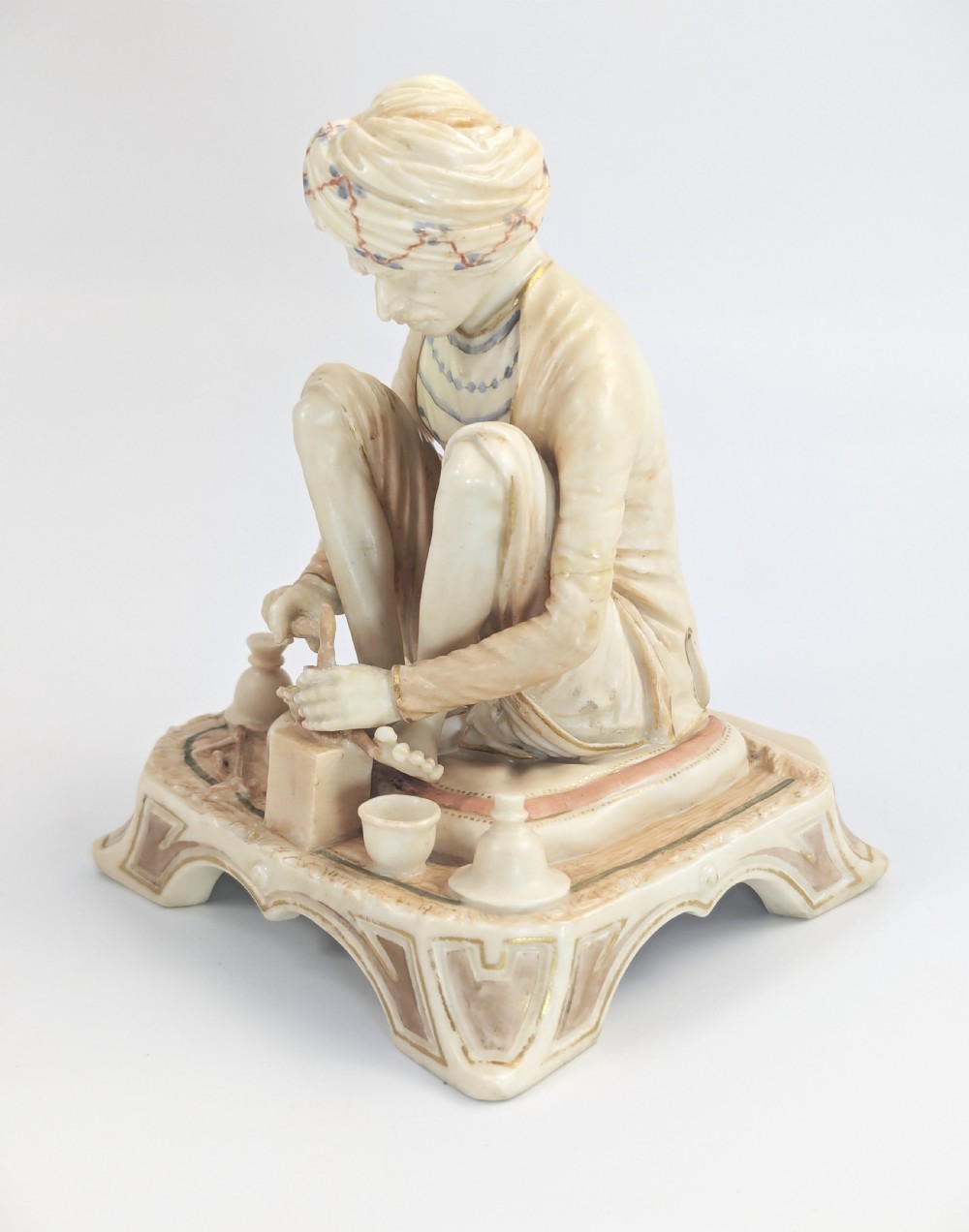 A late nineteenth-century Royal Worcester blush ivory porcelain figurine depicting a seated Indian craftsman working on an ornate pedestal base. View II