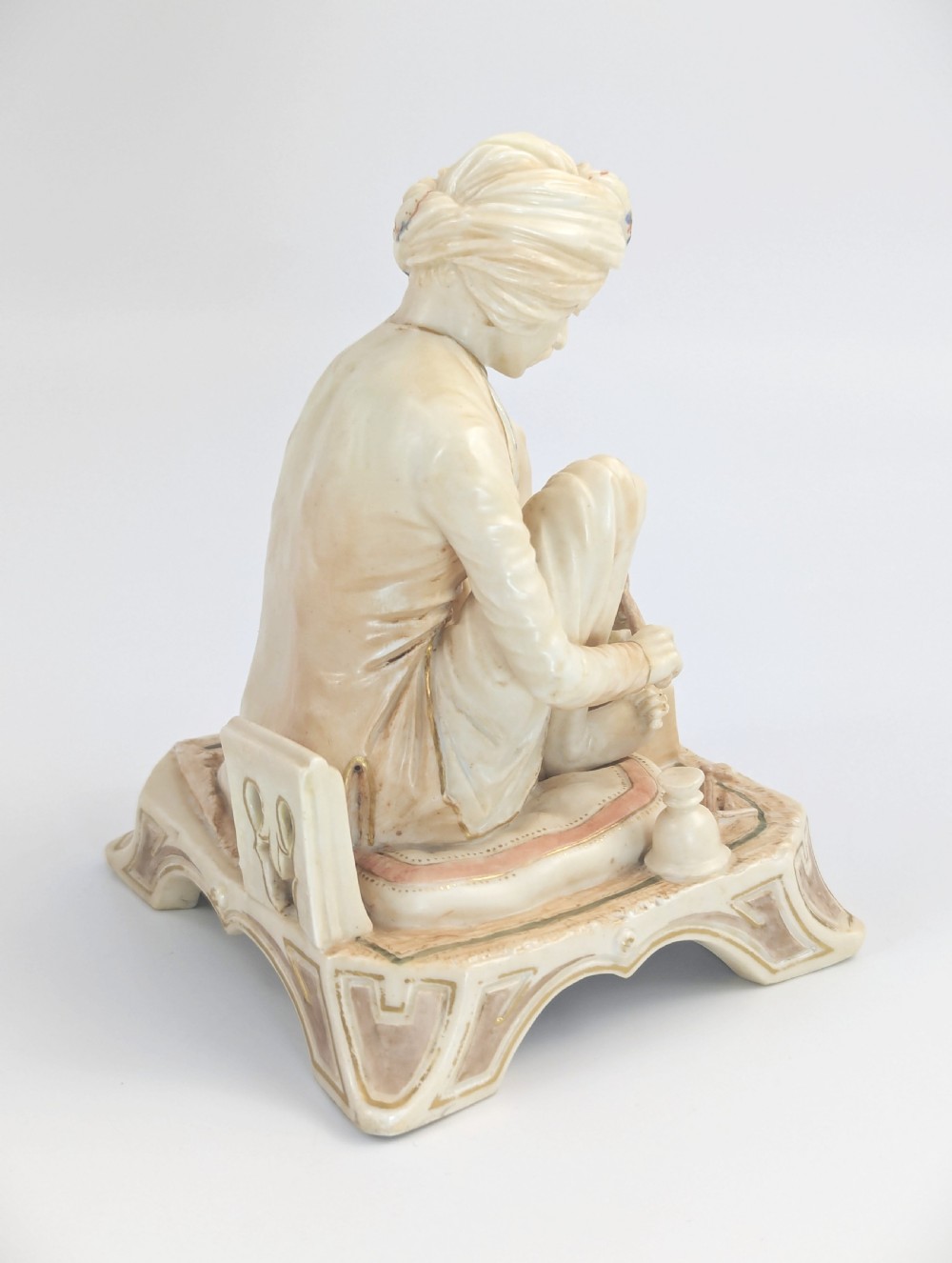 A late nineteenth-century Royal Worcester blush ivory porcelain figurine depicting a seated Indian craftsman working on an ornate pedestal base. View V