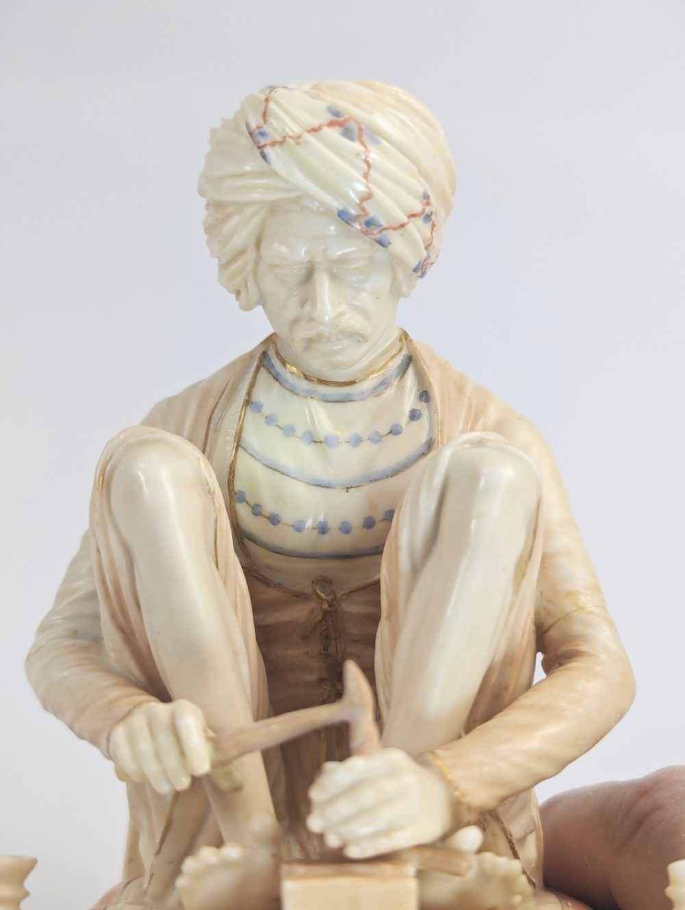 A late nineteenth-century Royal Worcester blush ivory porcelain figurine depicting a seated Indian craftsman working on an ornate pedestal base. View VIII