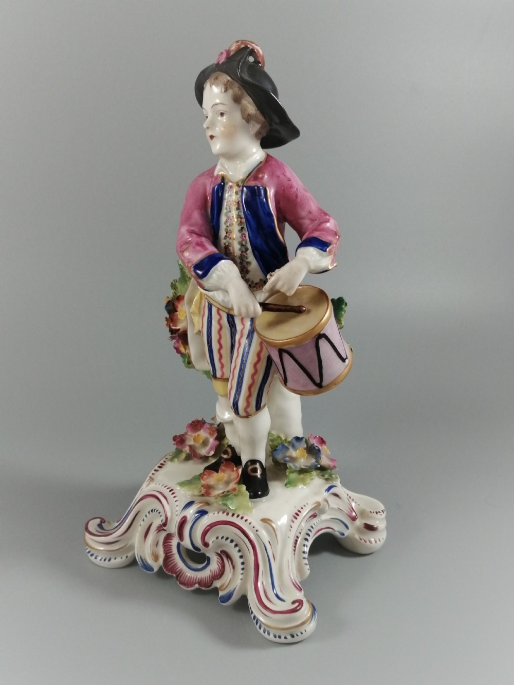 An 18th-century Bow porcelain figurine depicting a boy drummer wearing a pink coat, blue waistcoat, and tricorn hat on a Rococo scroll base with floral encrustations. View II