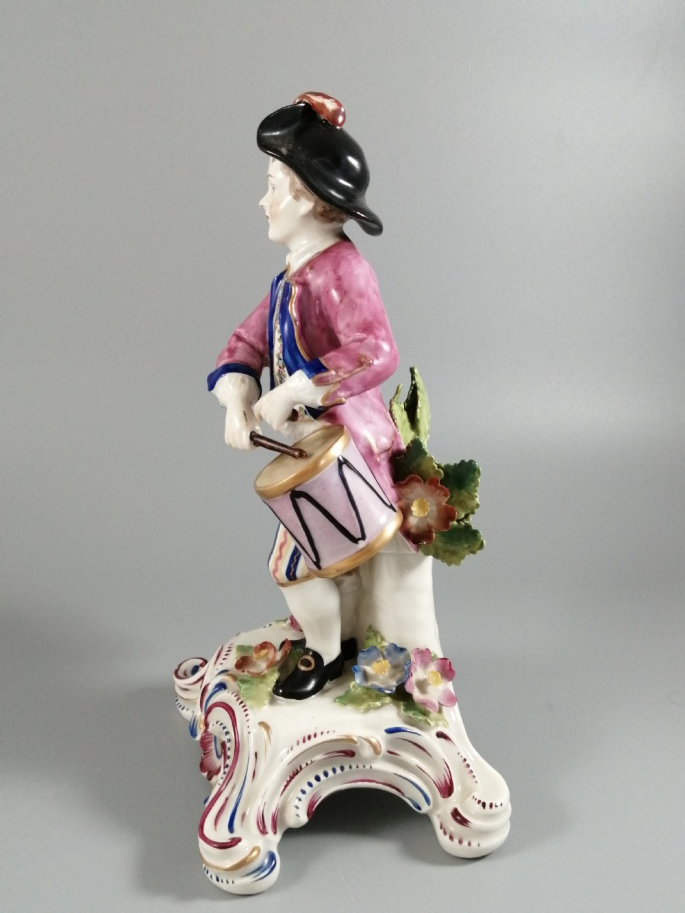 An 18th-century Bow porcelain figurine depicting a boy drummer wearing a pink coat, blue waistcoat, and tricorn hat on a Rococo scroll base with floral encrustations. View III