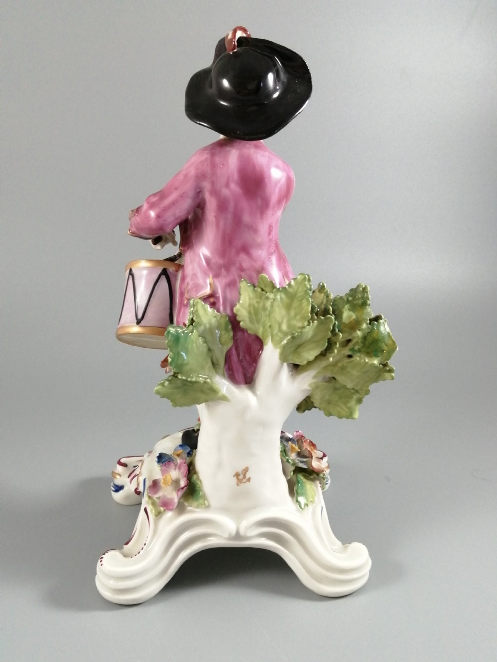 An 18th-century Bow porcelain figurine depicting a boy drummer wearing a pink coat, blue waistcoat, and tricorn hat on a Rococo scroll base with floral encrustations. View IV