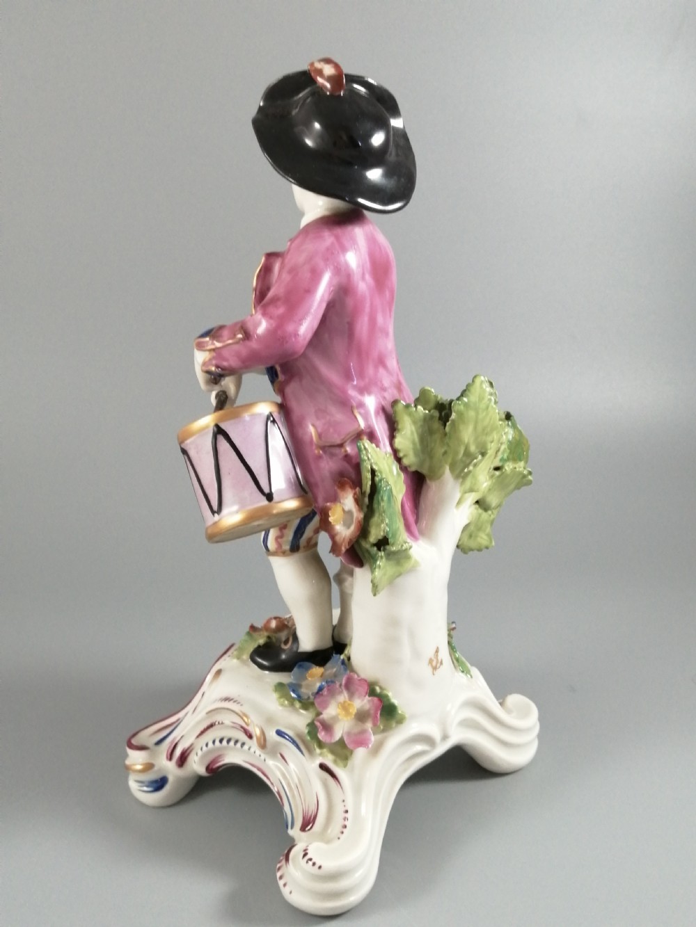 An 18th-century Bow porcelain figurine depicting a boy drummer wearing a pink coat, blue waistcoat, and tricorn hat on a Rococo scroll base with floral encrustations. View V