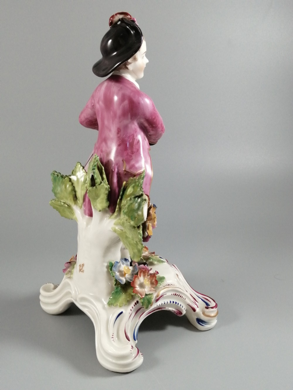An 18th-century Bow porcelain figurine depicting a boy drummer wearing a pink coat, blue waistcoat, and tricorn hat on a Rococo scroll base with floral encrustations. View VII