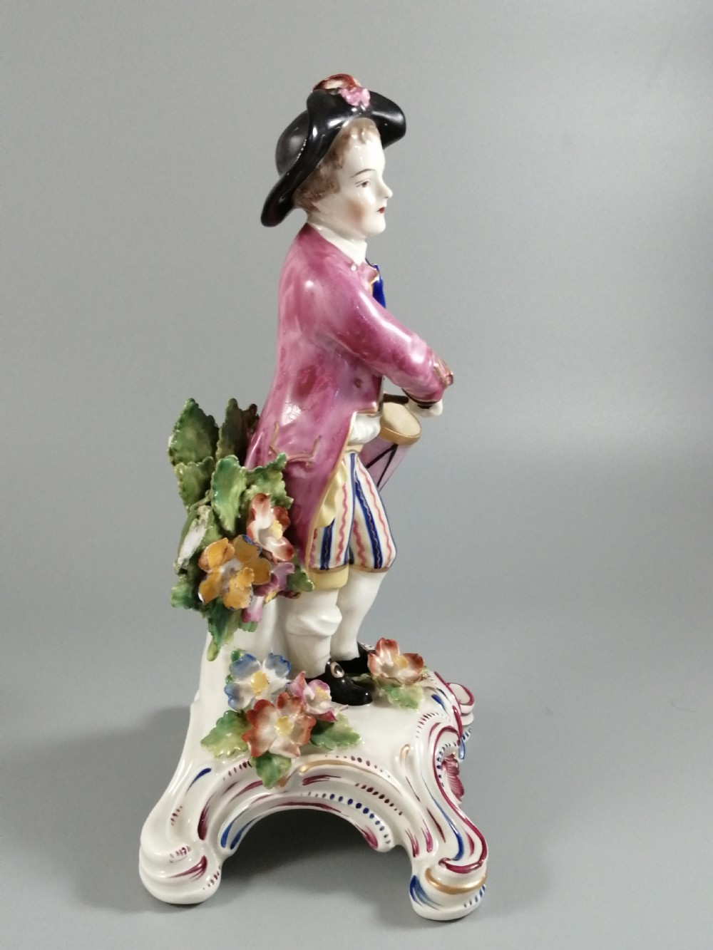 An 18th-century Bow porcelain figurine depicting a boy drummer wearing a pink coat, blue waistcoat, and tricorn hat on a Rococo scroll base with floral encrustations. View VIII