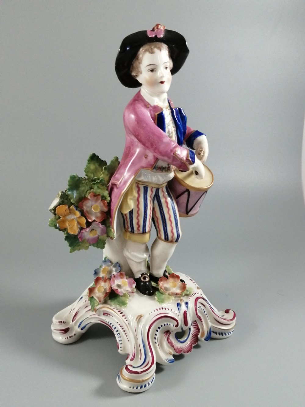 An 18th-century Bow porcelain figurine depicting a boy drummer wearing a pink coat, blue waistcoat, and tricorn hat on a Rococo scroll base with floral encrustations. View IX