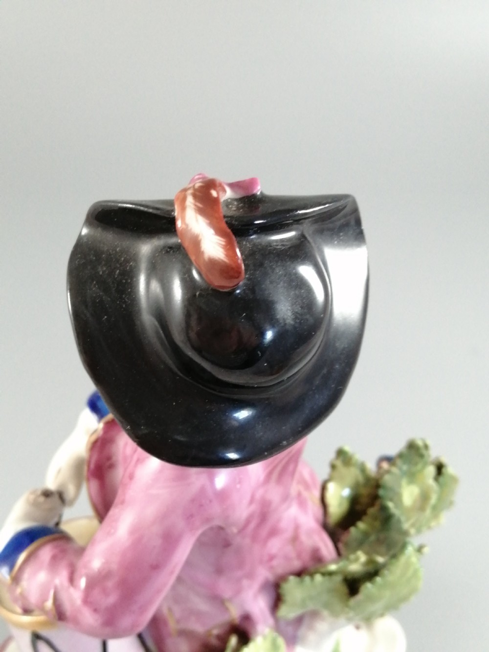 An 18th-century Bow porcelain figurine depicting a boy drummer wearing a pink coat, blue waistcoat, and tricorn hat on a Rococo scroll base with floral encrustations. View X