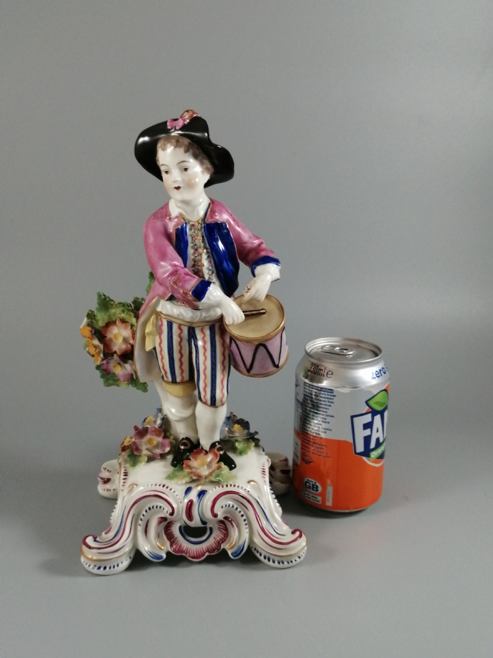 An 18th-century Bow porcelain figurine depicting a boy drummer wearing a pink coat, blue waistcoat, and tricorn hat on a Rococo scroll base with floral encrustations. View XII