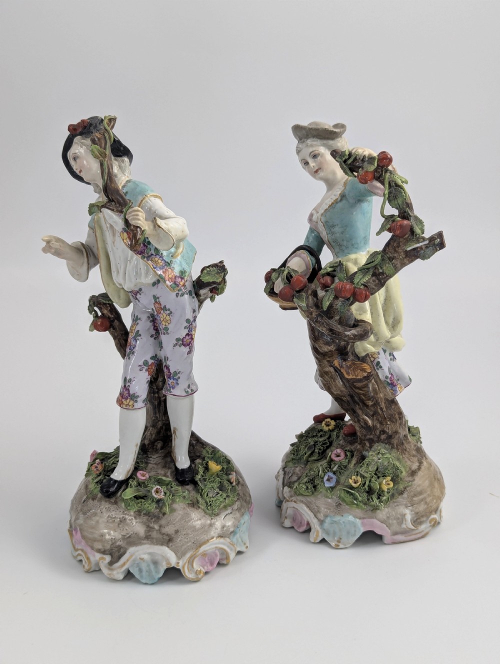 A pair of 18th-century German porcelain figurines depicting a male and female gardener standing beside fruiting trees on Rococo scrollwork bases. View II