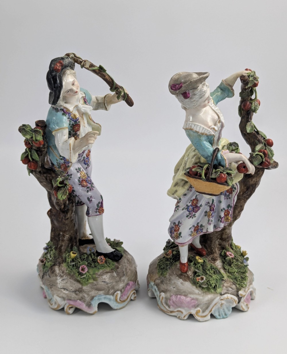 A pair of 18th-century German porcelain figurines depicting a male and female gardener standing beside fruiting trees on Rococo scrollwork bases. View V