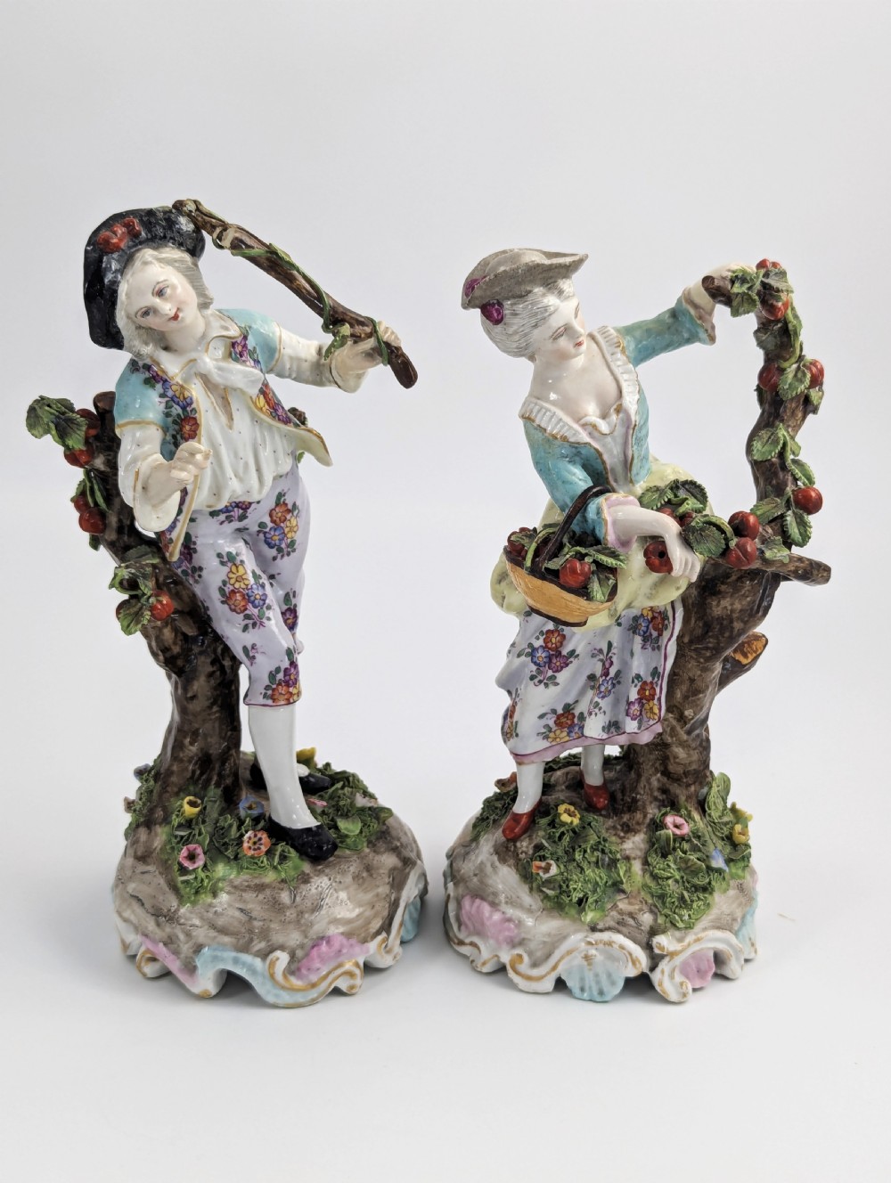 A pair of 18th-century German porcelain figurines depicting a male and female gardener standing beside fruiting trees on Rococo scrollwork bases. View VI