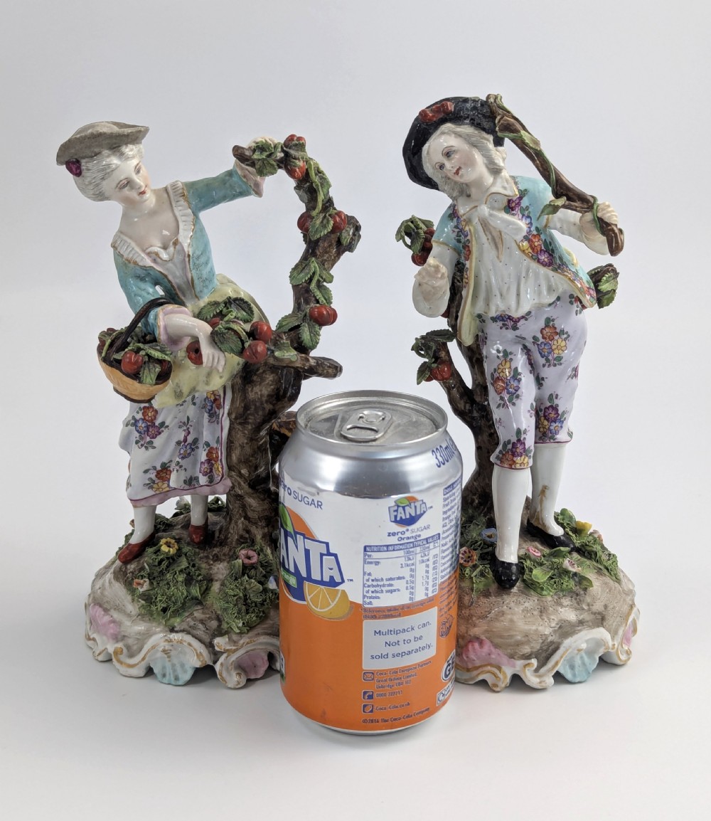 A pair of 18th-century German porcelain figurines depicting a male and female gardener standing beside fruiting trees on Rococo scrollwork bases. View IX