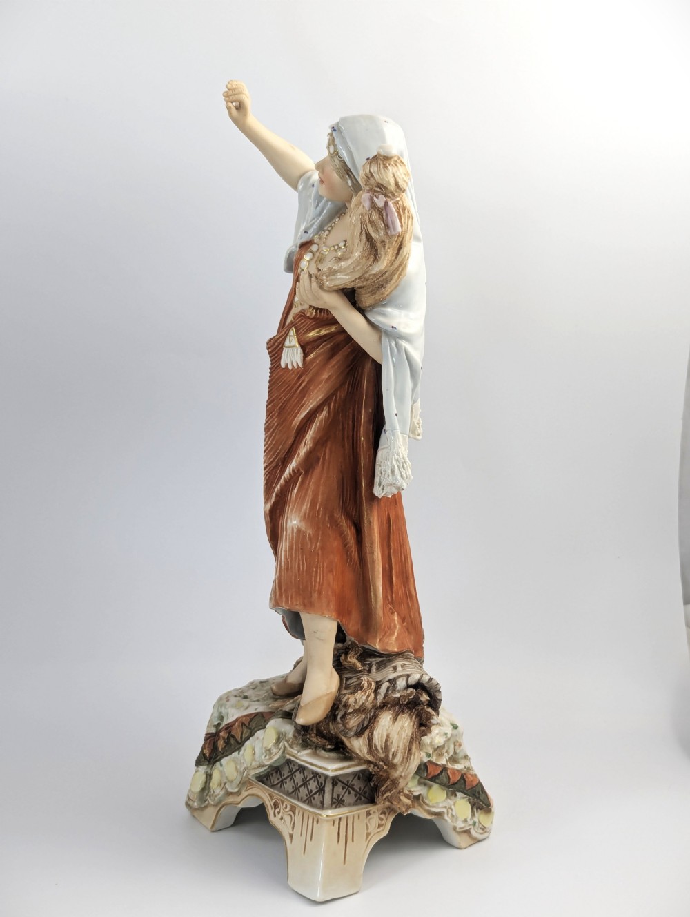 An antique Royal Dux porcelain figurine depicting a standing young woman wearing draped terracotta and cream robes on a decorative rocky pedestal. View III