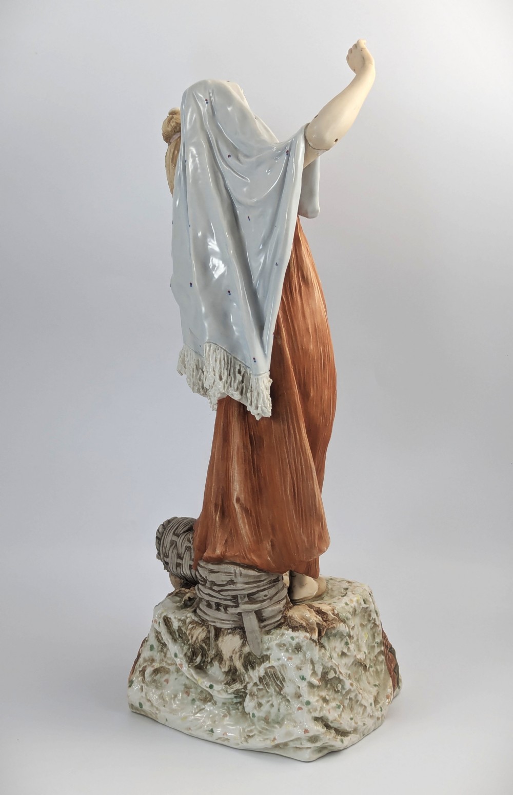 An antique Royal Dux porcelain figurine depicting a standing young woman wearing draped terracotta and cream robes on a decorative rocky pedestal. View VI