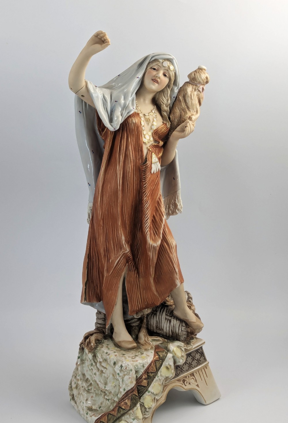 An antique Royal Dux porcelain figurine depicting a standing young woman wearing draped terracotta and cream robes on a decorative rocky pedestal. View IX