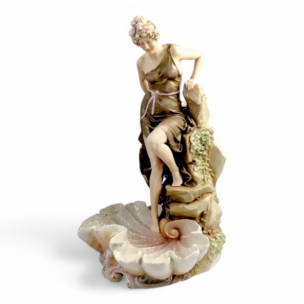 An Art Nouveau Royal Dux porcelain centrepiece depicting a draped female bather standing atop rockwork above a large shell-shaped basin.