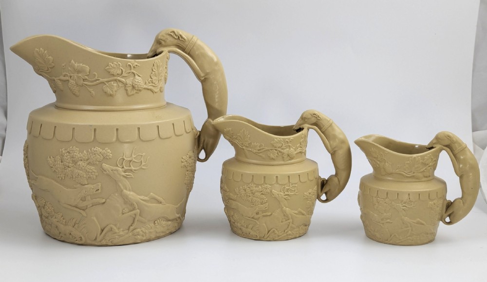 A graduated set of three buff yellow Regency ceramic pitchers featuring relief-moulded stag hunting scenes and sculpted hound handles. View II