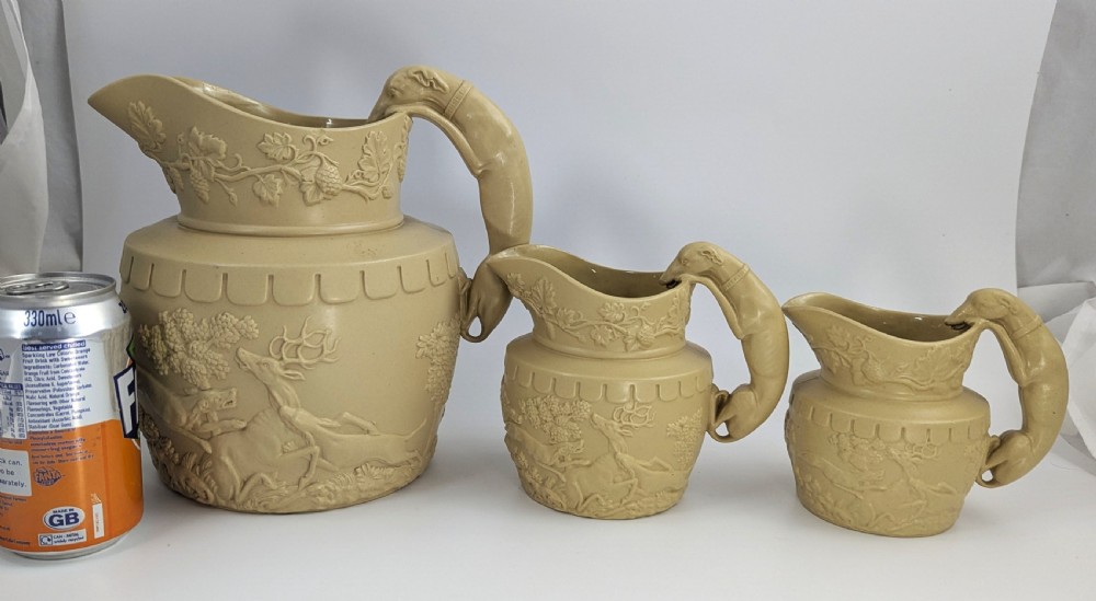 A graduated set of three buff yellow Regency ceramic pitchers featuring relief-moulded stag hunting scenes and sculpted hound handles. View IX