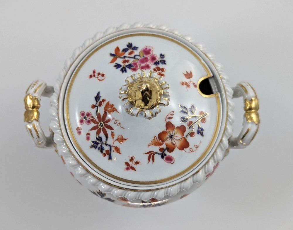 A white Regency porcelain sauce tureen decorated with orange and blue floral Imari motifs, featuring twin gilded handles, a square pedestal base, and a domed lid with a gilded finial. View IX