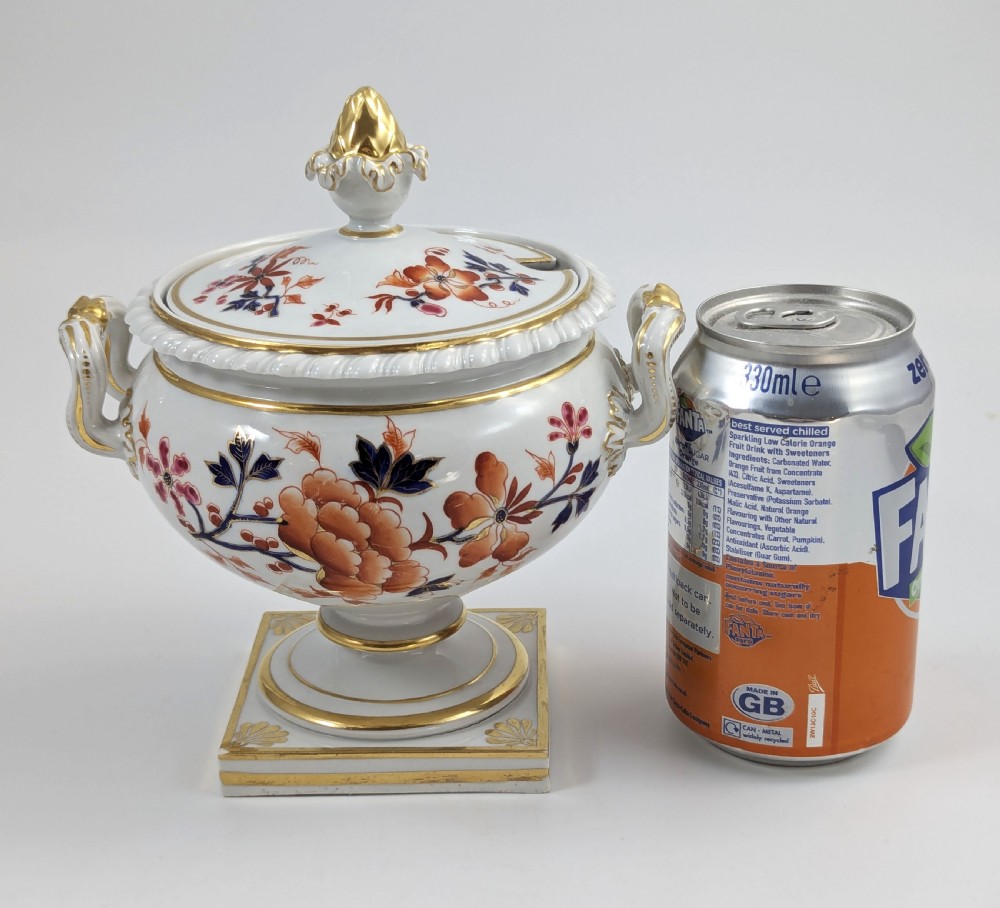 A white Regency porcelain sauce tureen decorated with orange and blue floral Imari motifs, featuring twin gilded handles, a square pedestal base, and a domed lid with a gilded finial. View X