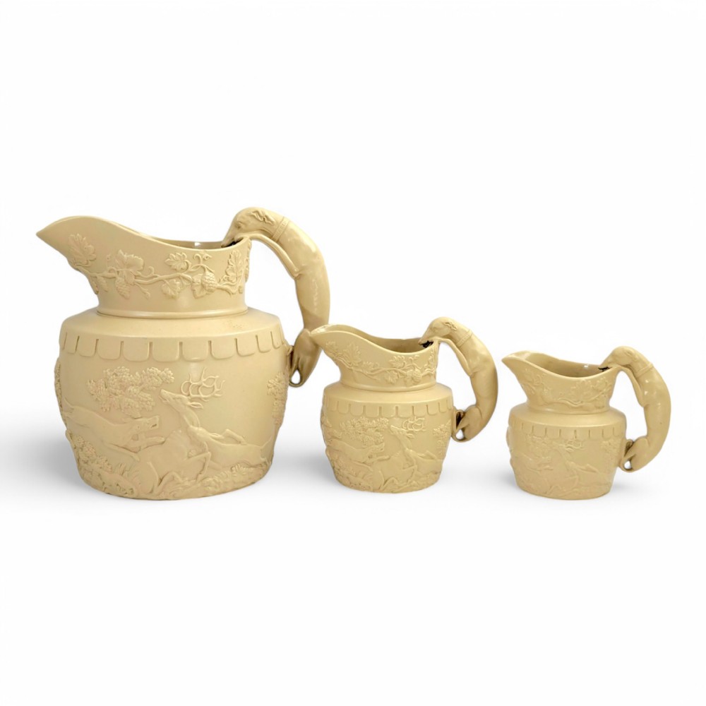 A graduated set of three buff yellow Regency ceramic pitchers featuring relief-moulded stag hunting scenes and sculpted hound handles.