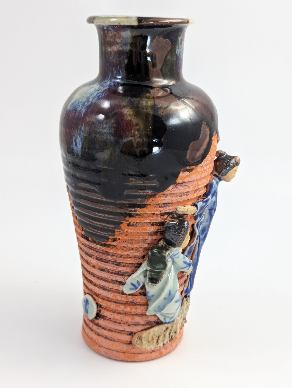 A Japanese Sumida Gawa pottery vase featuring an oxblood flambe glazed neck and applied relief figures on a ribbed terracotta body. View V