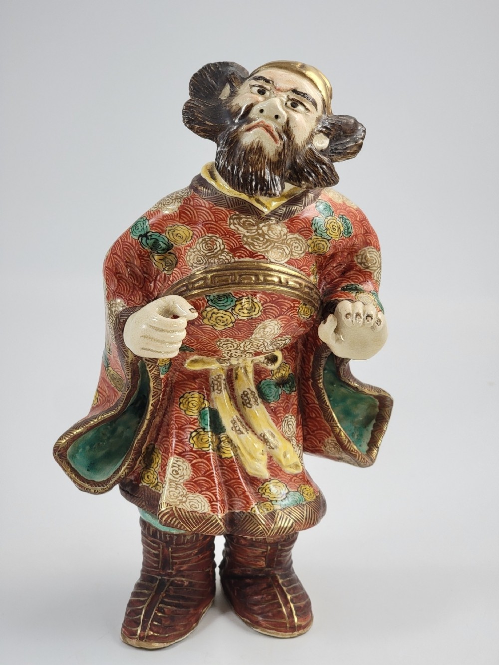 A Japanese Kutani porcelain figurine depicting a bearded male figure in a red and gold patterned robe with raised moriage enamel detailing.