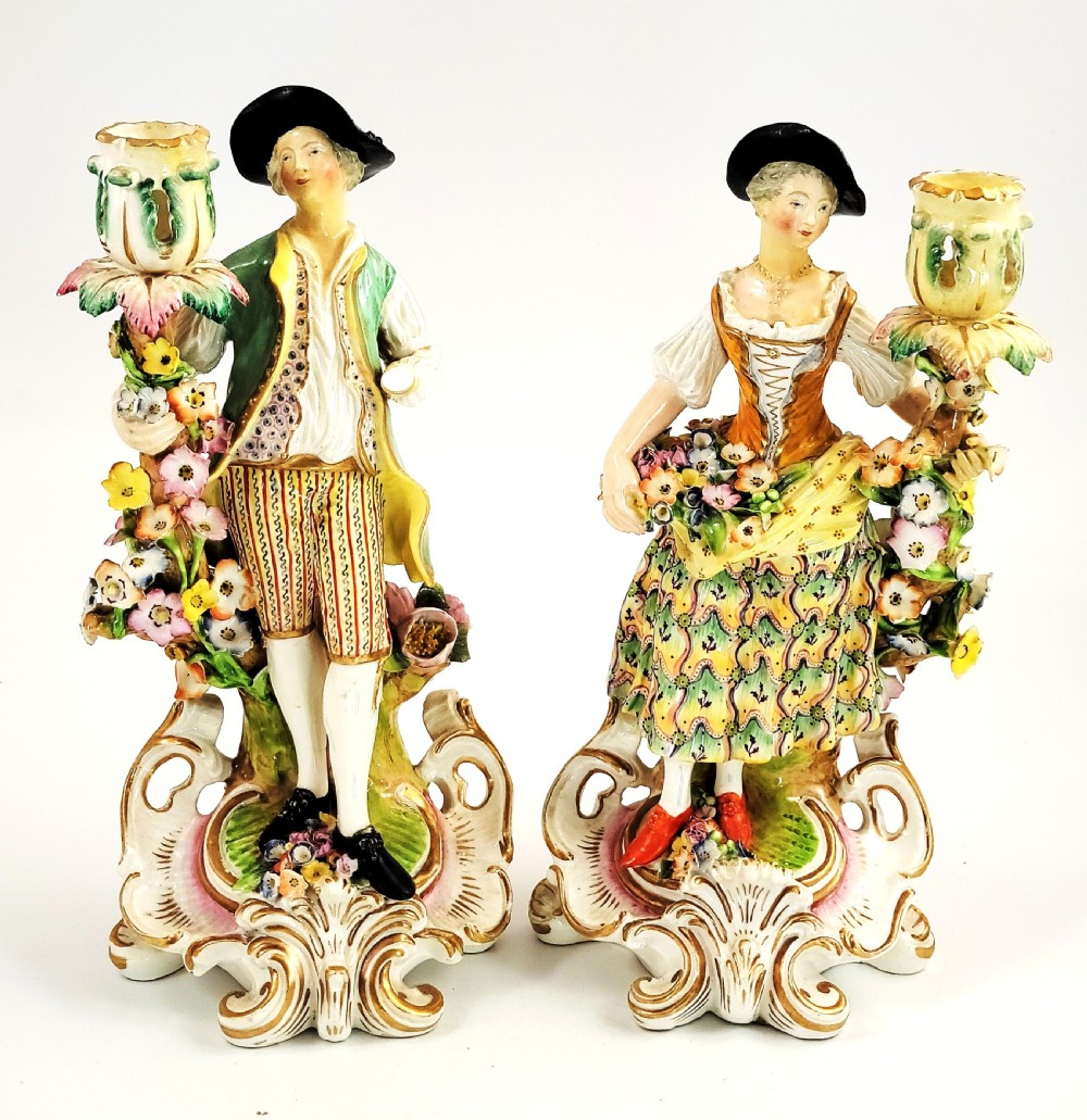 A pair of 18th-century Georgian Derby soft-paste porcelain figural candlesticks depicting a male and female figure in period dress beside candle sconces adorned with colourful flowers and gilt Rococo scrollwork.