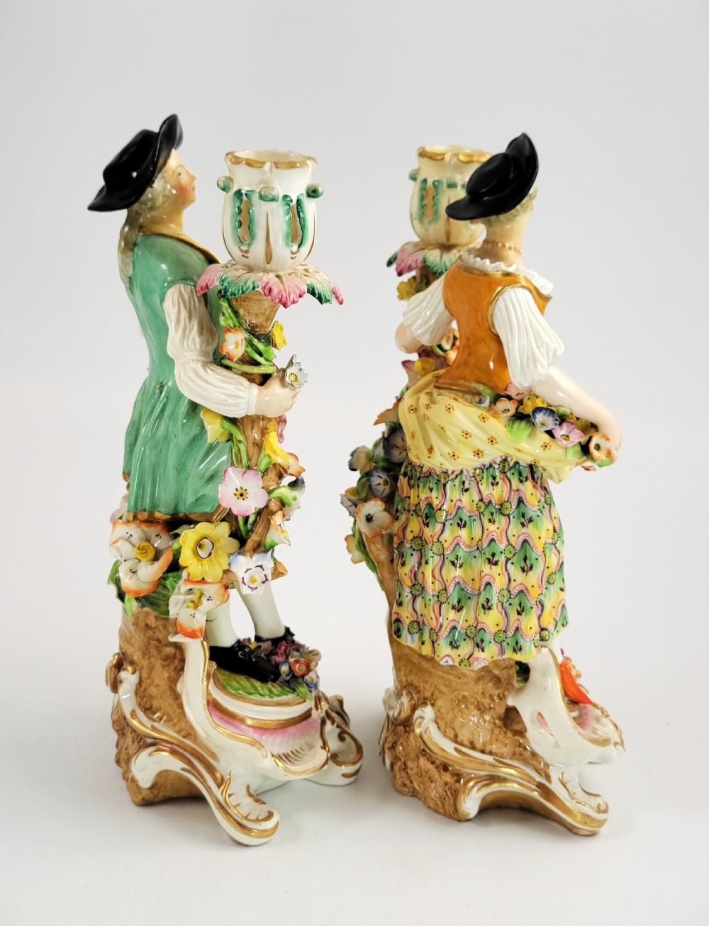 A pair of 18th-century Georgian Derby soft-paste porcelain figural candlesticks depicting a male and female figure in period dress beside candle sconces adorned with colourful flowers and gilt Rococo scrollwork. View V