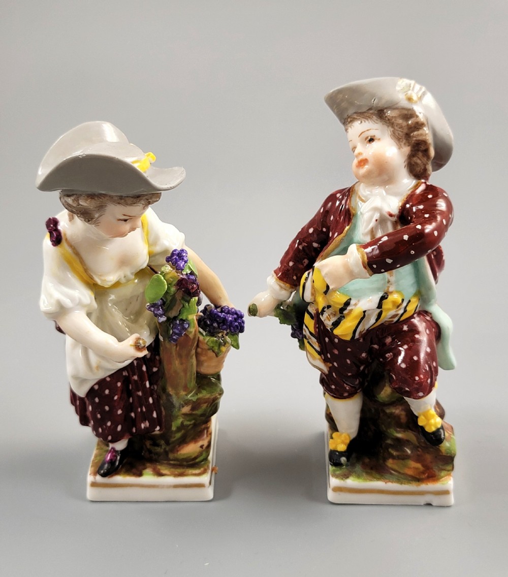 A pair of 19th-century Dresden porcelain figurines depicting a male and female grape picker in traditional attire holding baskets of purple grapes.