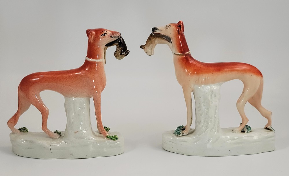 A pair of Victorian Staffordshire earthenware greyhound figurines standing on white bases with caught hares held in their mouths.