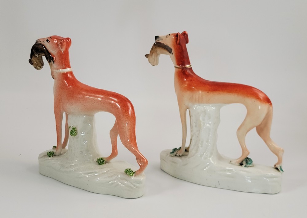 A pair of Victorian Staffordshire earthenware greyhound figurines standing on white bases with caught hares held in their mouths. View II
