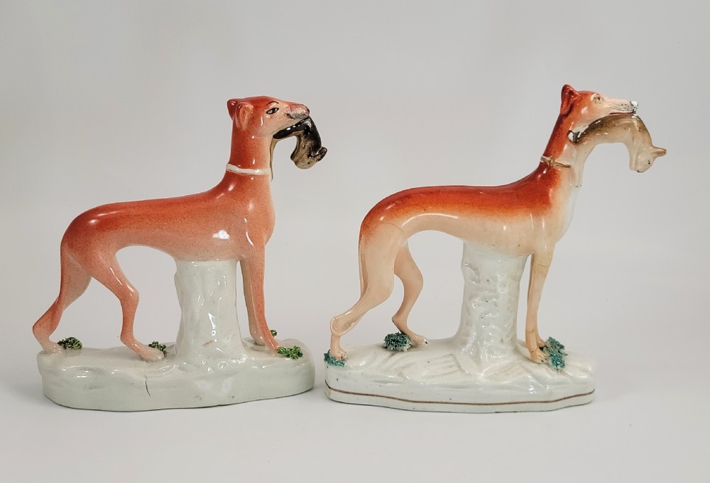 A pair of Victorian Staffordshire earthenware greyhound figurines standing on white bases with caught hares held in their mouths. View IV
