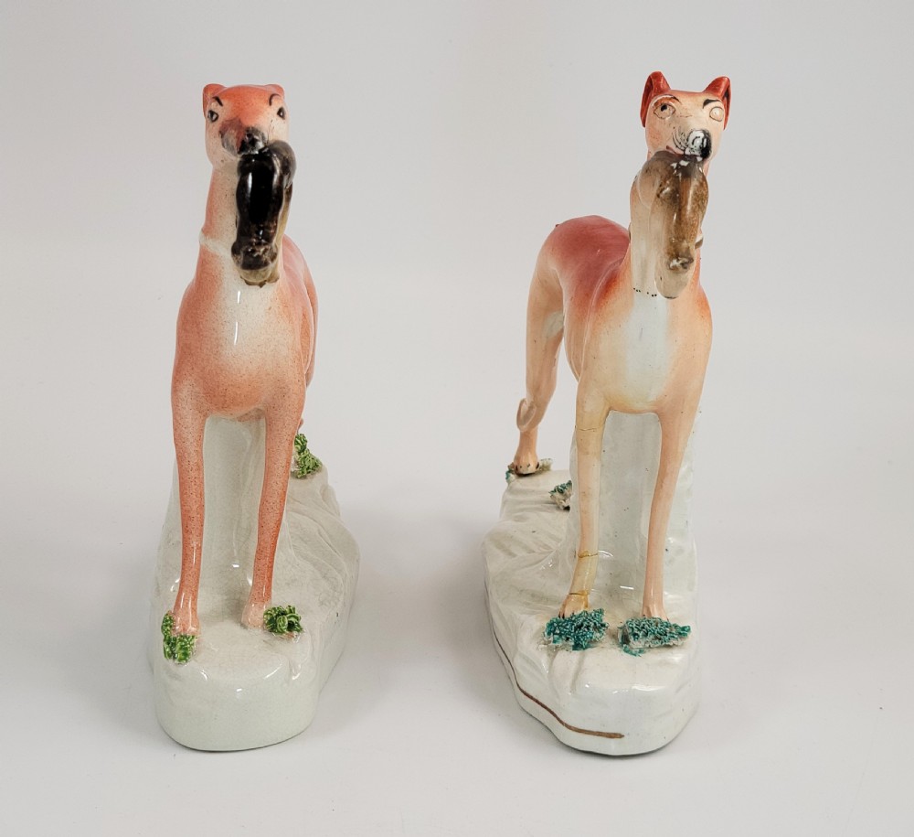 A pair of Victorian Staffordshire earthenware greyhound figurines standing on white bases with caught hares held in their mouths. View V