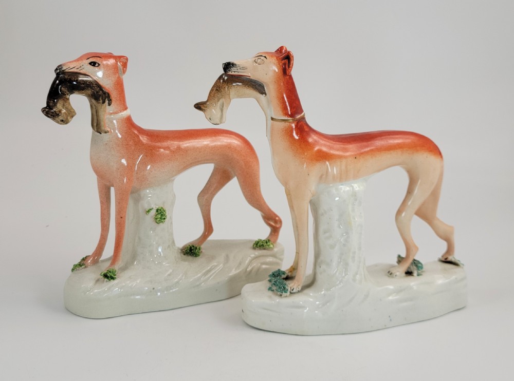 A pair of Victorian Staffordshire earthenware greyhound figurines standing on white bases with caught hares held in their mouths. View VI
