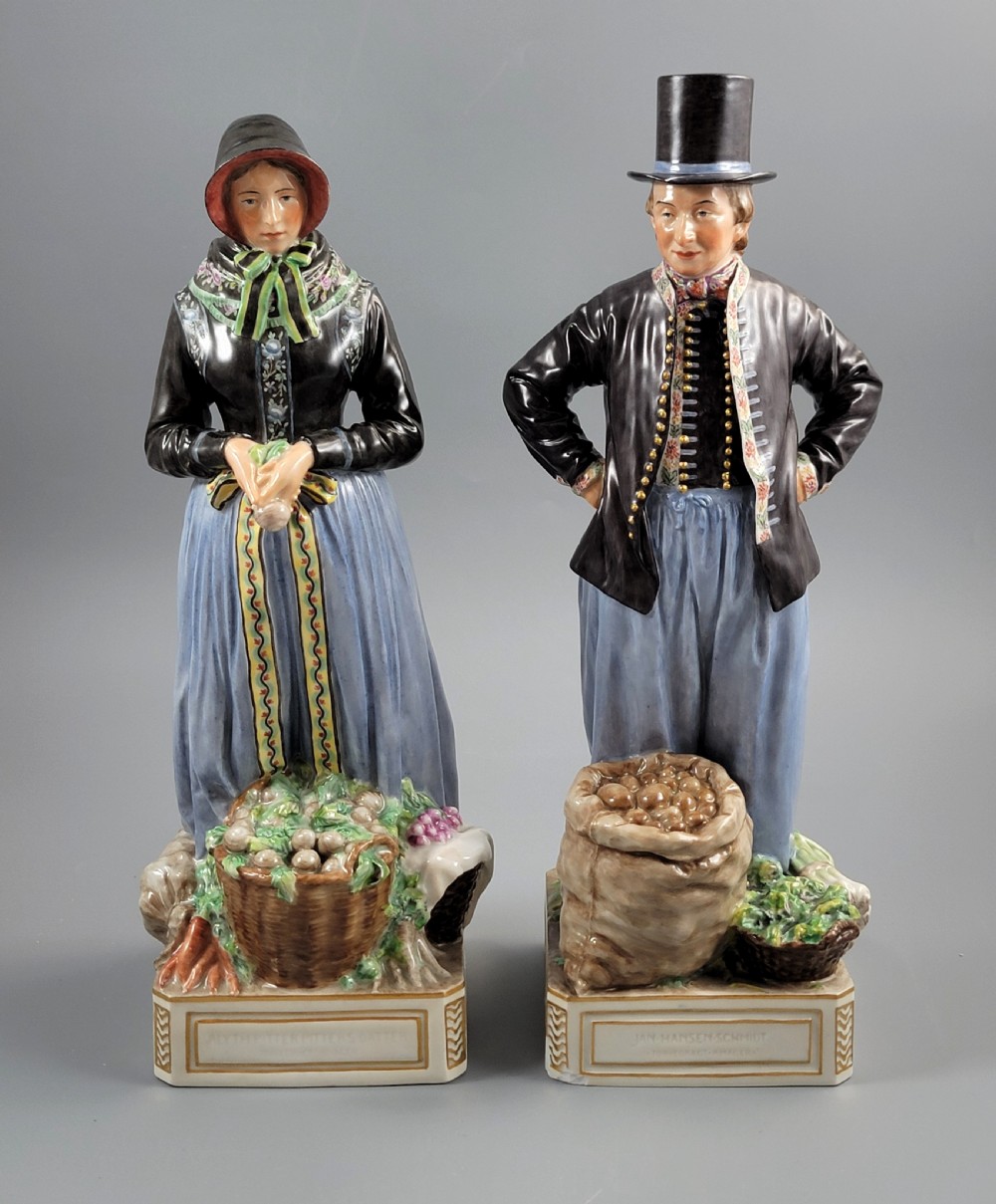 A pair of Royal Copenhagen glazed porcelain figurines depicting a man and woman dressed in traditional Danish Amager market costumes alongside baskets of produce.