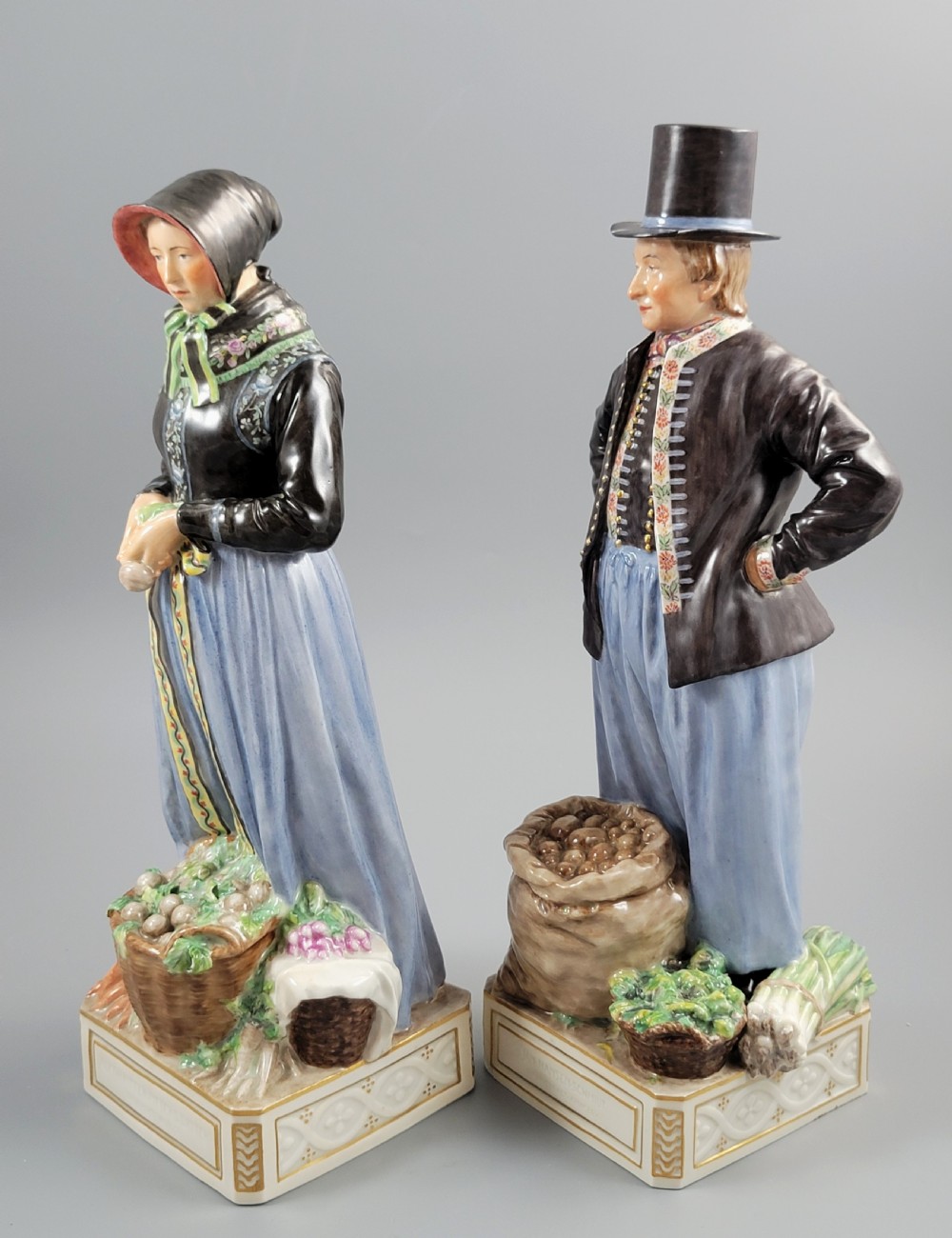 A pair of Royal Copenhagen glazed porcelain figurines depicting a man and woman dressed in traditional Danish Amager market costumes alongside baskets of produce. View II