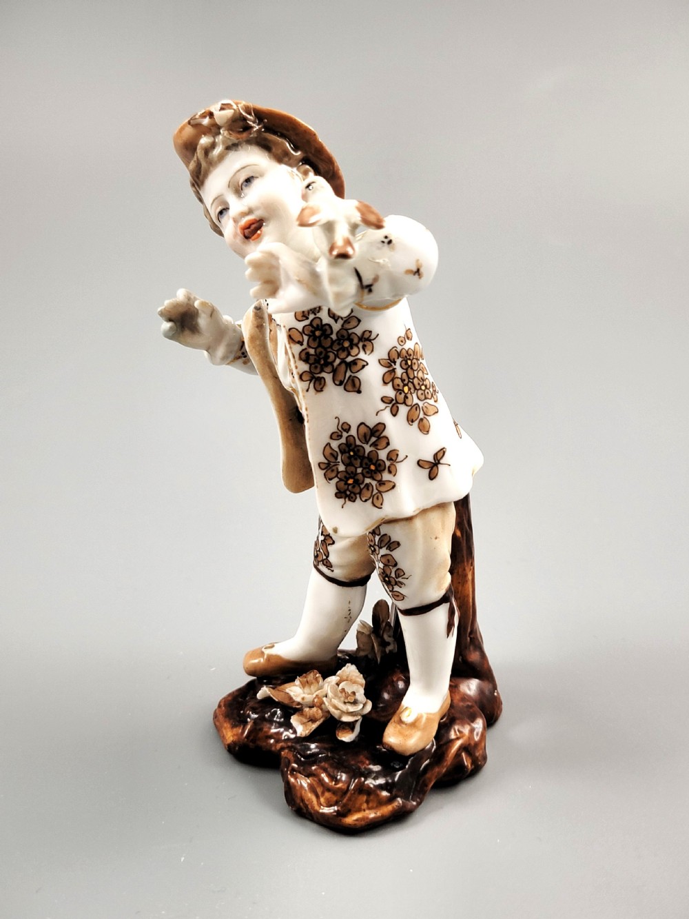 An antique German porcelain figurine depicting a boy wearing a floral coat and holding a small bird on his hand. View II