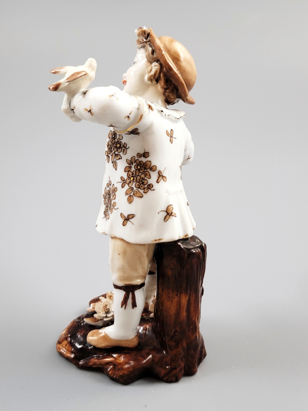 An antique German porcelain figurine depicting a boy wearing a floral coat and holding a small bird on his hand. View III