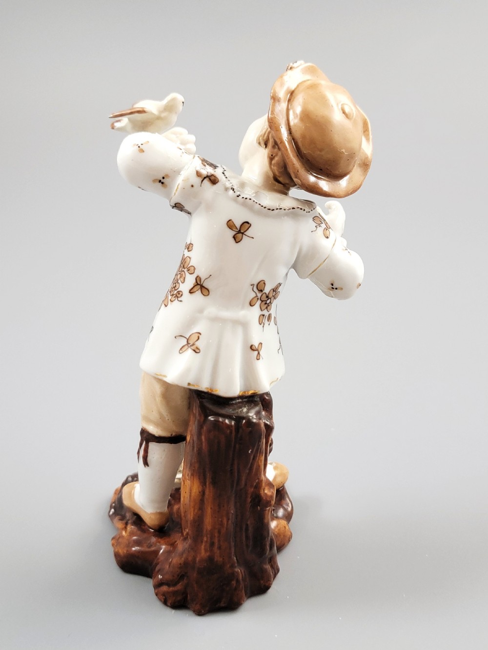 An antique German porcelain figurine depicting a boy wearing a floral coat and holding a small bird on his hand. View IV