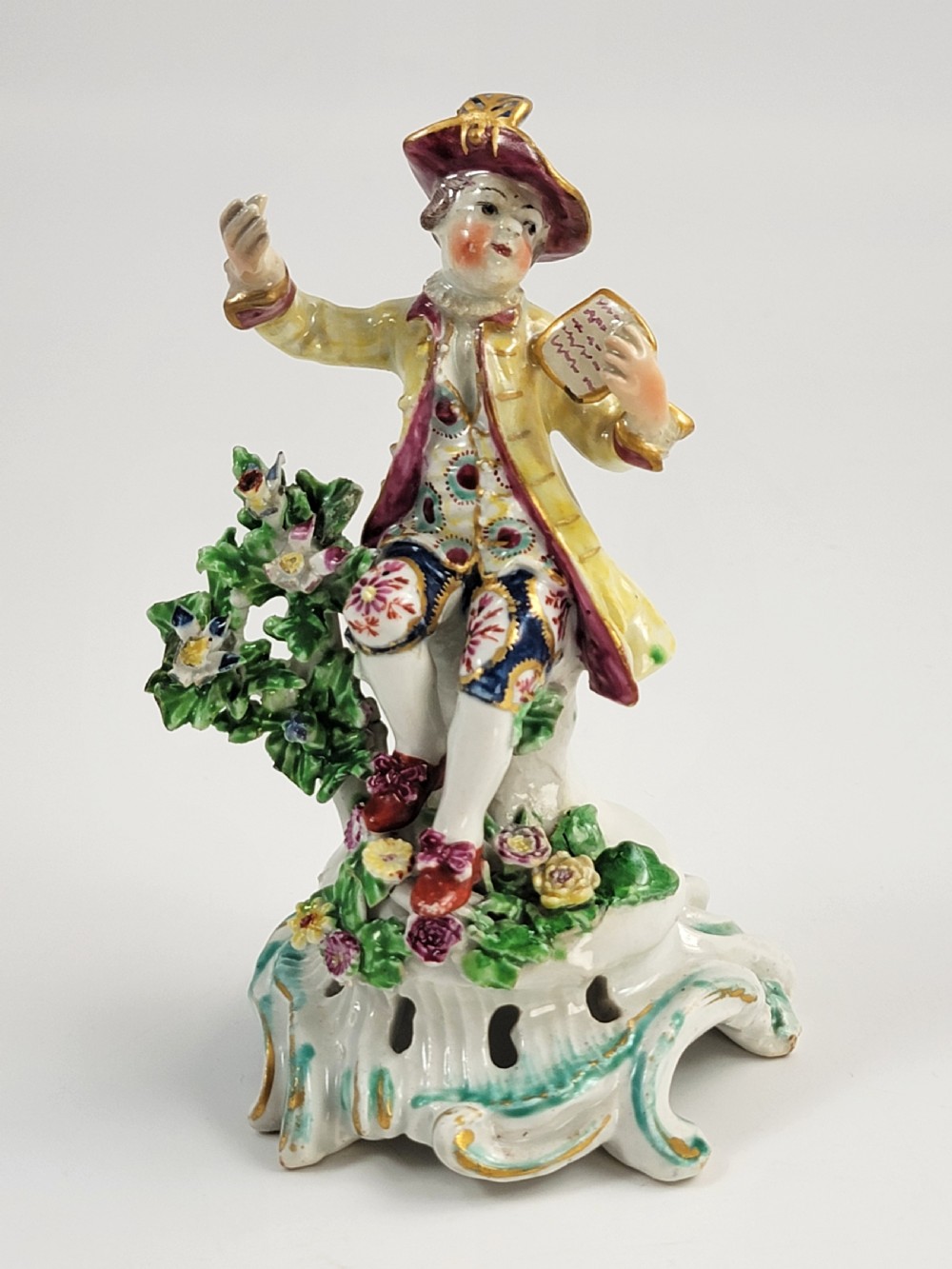 An eighteenth-century Bow soft-paste porcelain figurine depicting a seated boy singer holding sheet music atop a pierced scrollwork rococo base with applied floral bocage.