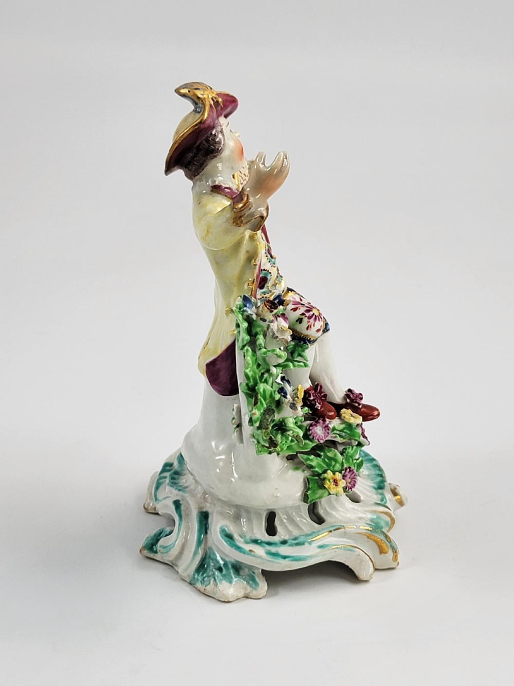 An eighteenth-century Bow soft-paste porcelain figurine depicting a seated boy singer holding sheet music atop a pierced scrollwork rococo base with applied floral bocage. View VI