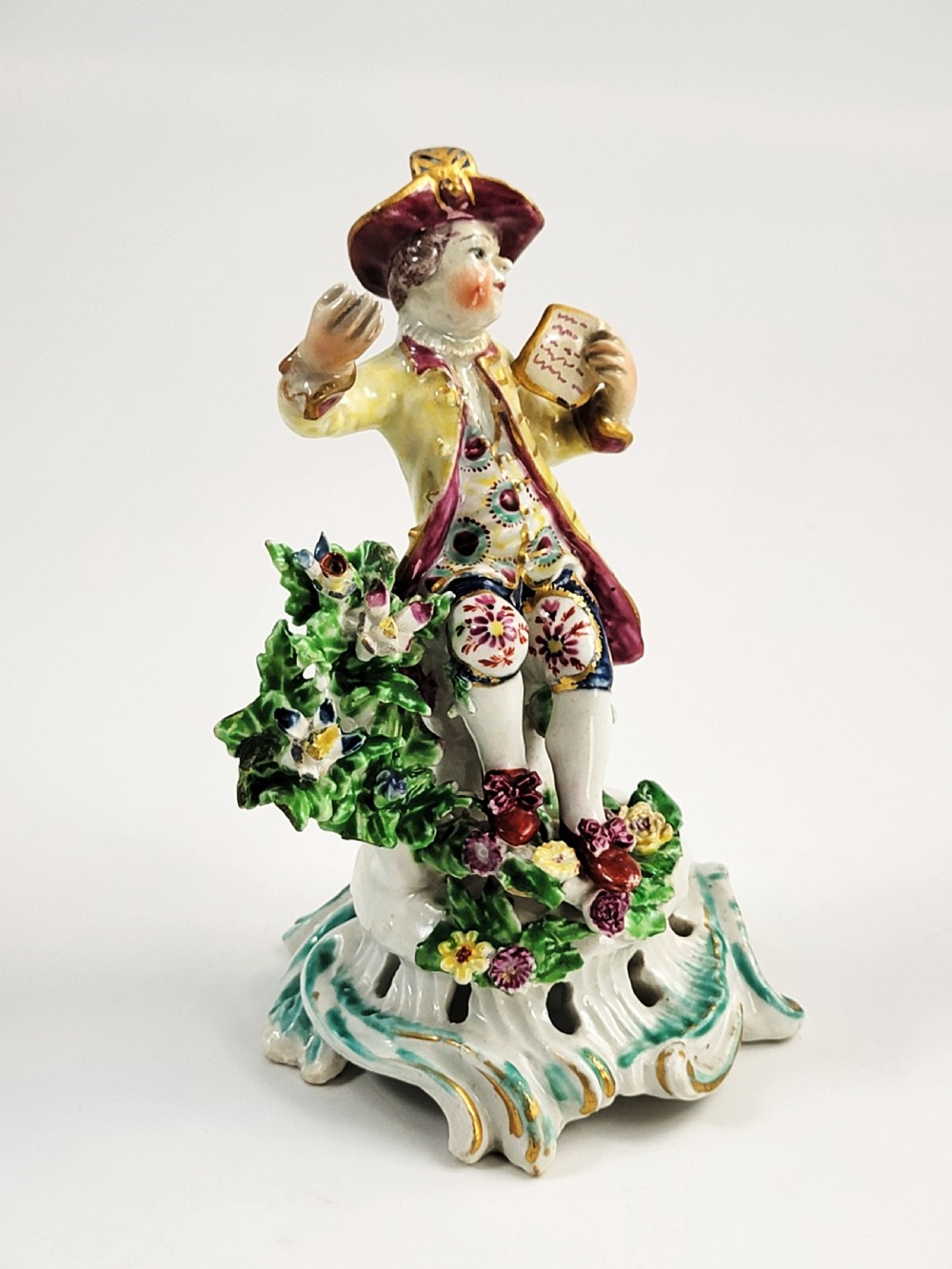 An eighteenth-century Bow soft-paste porcelain figurine depicting a seated boy singer holding sheet music atop a pierced scrollwork rococo base with applied floral bocage. View VII