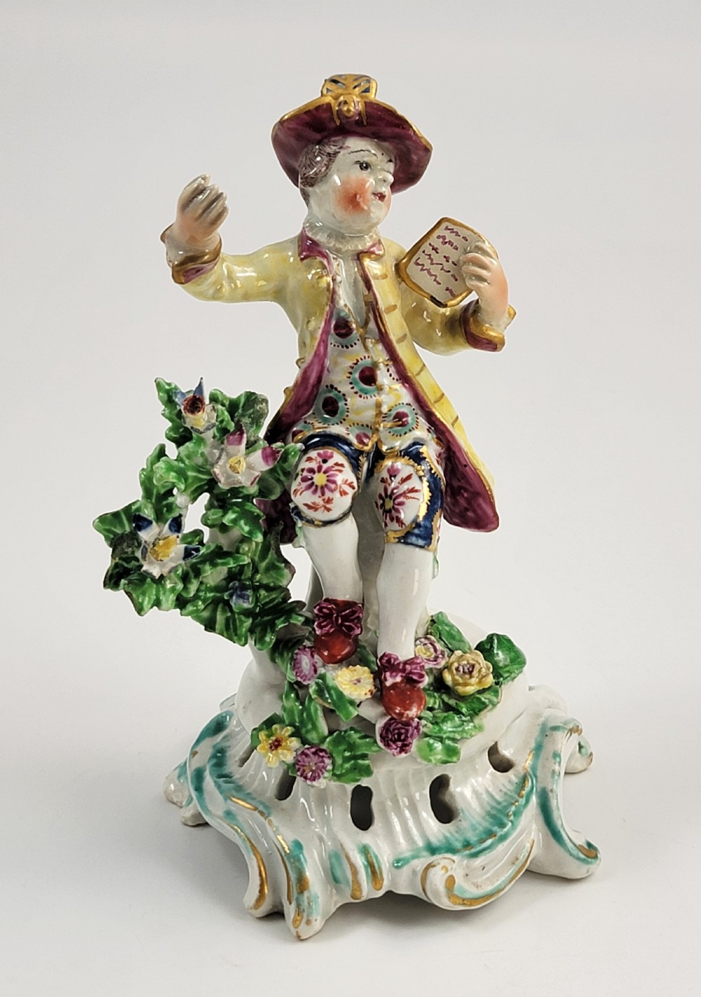 An eighteenth-century Bow soft-paste porcelain figurine depicting a seated boy singer holding sheet music atop a pierced scrollwork rococo base with applied floral bocage. View VIII