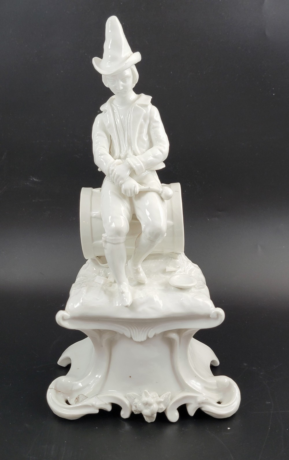 An 18th-century monochrome white glazed porcelain figurine of a male figure seated on a drum atop a Rococo scrollwork base. View IV