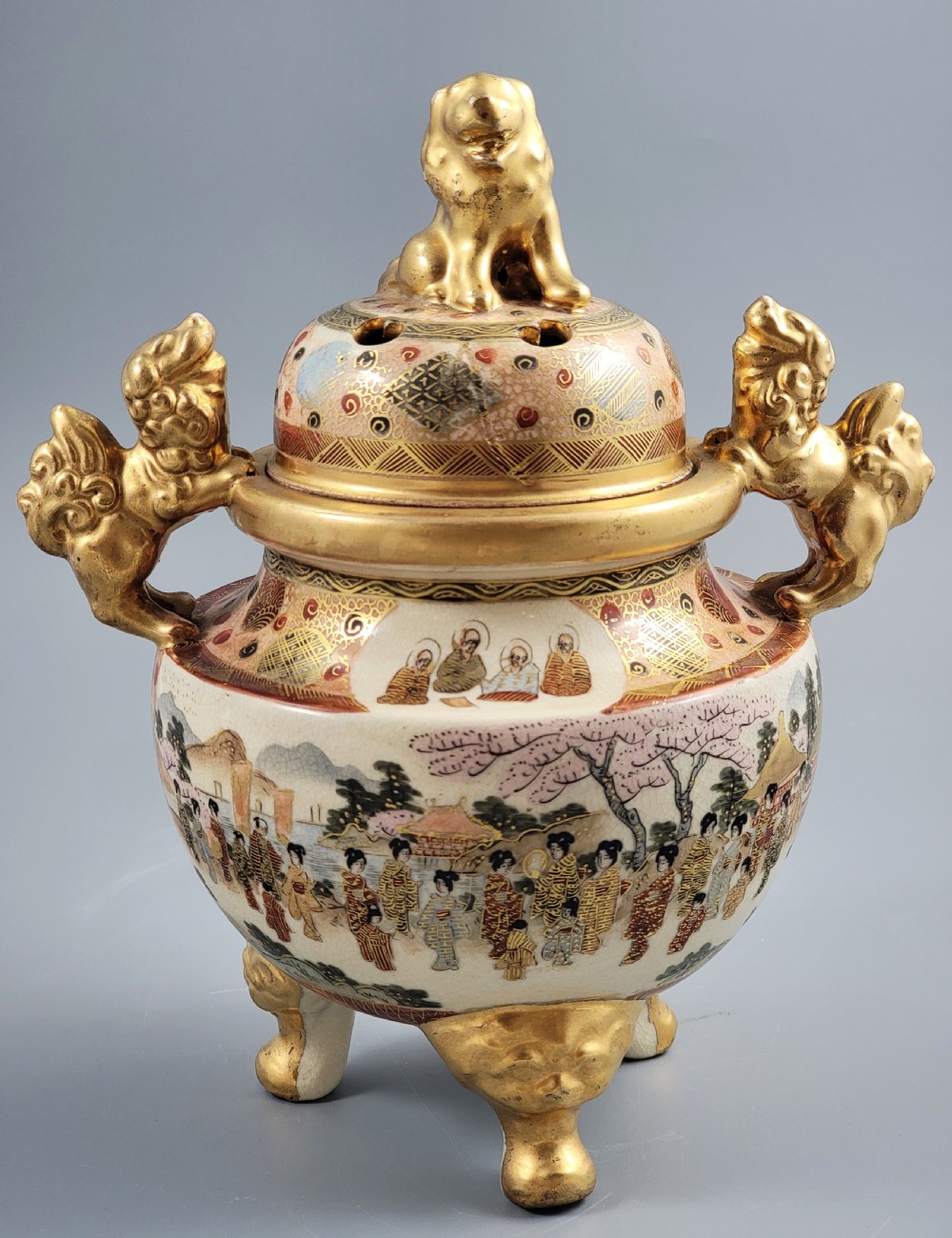 A Japanese Meiji period Satsuma earthenware incense burner featuring gilt shishi handles and finial with detailed hand-painted figures over a crackle glaze.