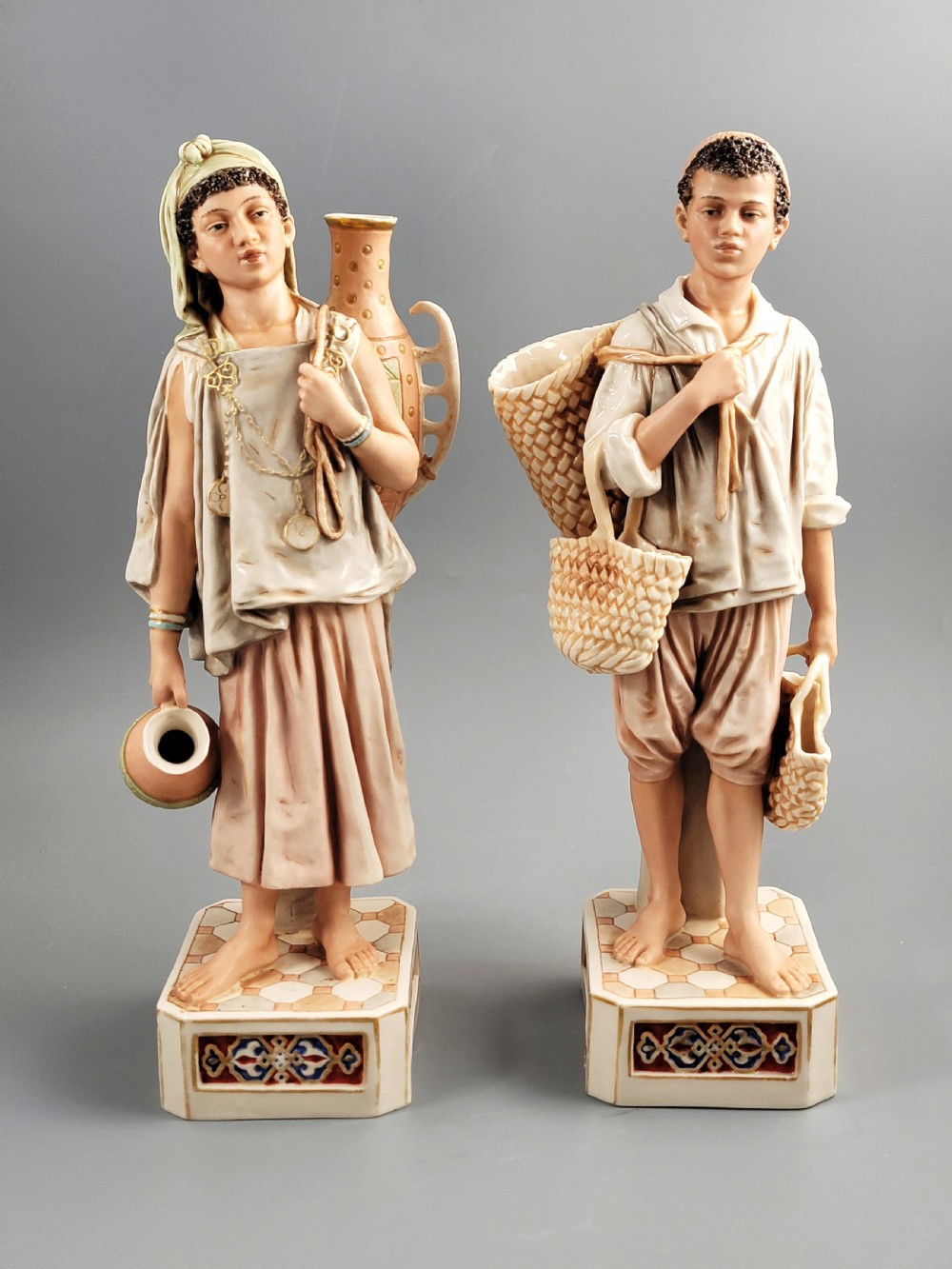 A pair of late 19th-century Royal Dux porcelain figurines depicting a male and female water bearer in draped attire standing on patterned pedestal bases.